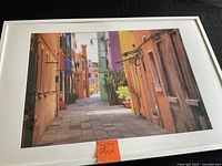 Front view of framed photograph showing colourful narrow street, white frame, size reference label