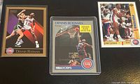 Three Dennis Rodman Detroit Pistons trading cards from SkyBox, NBA Hoops, Upper Deck laid on black surface