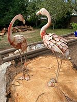 Two painted metal flamingo sculptures standing on tree-stump base