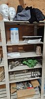 Shelving unit containing listed items: two mannequin heads, folded textiles, plastic container, books, miniature wheelbarrow, artificial foliage, metal wall rack, garland in box, woven basket