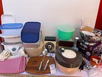 Wide view of entire lot including boxed carafe, scale, salad spinner, storage containers, steamer parts, utensils, linens