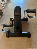 Front view of black under-desk pedal exerciser showing LCD, pedals, stabilizer bars and tension knob