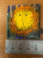 Front view of signed enamel-on-copper tile with ruler for scale