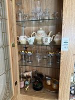 Full cabinet view showing glass stemware, silver/gold goblets, ceramic tea set, small bottles and pitchers