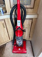 Full view of red Dirt Devil upright vacuum standing against cabinet