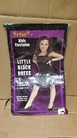 Front of sealed package showing Spirit Kids Costume Little Black Dress, model photo and size options