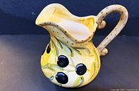 three-quarter view of ceramic pitcher showing spout, olive reliefs, yellow glaze