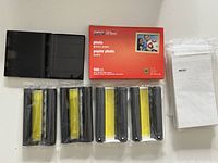 Lot overview showing five ink ribbon cartridges, Staples photo paper box, additional paper stack, and black case