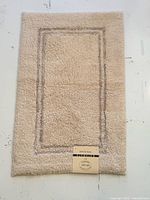 Full view of light yellow Alkaso bath mat with rectangular border and hang tag