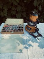 Router standing upright with power cord coiled, accessory rods adjacent, and open wooden case displaying multiple router bits