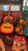 Guitar, violin in case, straps and instruction book on sofa