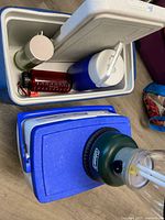 Open blue Coleman cooler with jug, thermos and bottles inside alongside second closed cooler