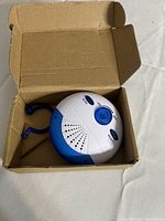 Detector in open cardboard box showing front with dial, buttons, speaker vents and blue hook antenna
