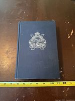 Front cover showing blue cloth boards and silver crest with measuring tape for scale