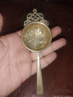 Front view of silver plated tea strainer showing bowl perforations, handle, and ornate crest
