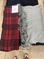 Red/black plaid shawl and light grey fringed shawl side by side on black surface