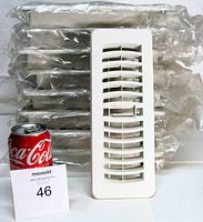 Stack of nine bagged white plastic floor registers with one unbagged register shown upright beside soda can for scale