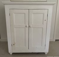 Front view of white two-door cabinet closed, showing paneled doors, knobs, bun feet, and overall appearance