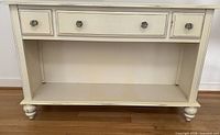 Full front view of console table showing open lower compartment, three drawer fronts, turned legs, ivory distressed paint