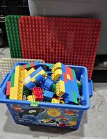 Blue bin filled with assorted Duplo bricks, red and green baseplates behind