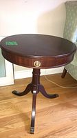 Full view of round pedestal side table showing top, drawer knob, pedestal, three legs