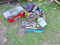 Overall view of toolbox, bolt cutter, weed puller, measuring jug and box of assorted hand tools on grass