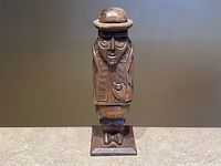 Front view of carved wooden figure showing facial features, bowler hat, jacket, hand on chest, square base