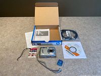 Lot overview showing camera, box, paperwork, software CD, sealed cable, SD card and wrist strap