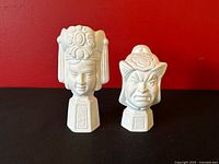 Front view of both bisque ceramic mask head sculptures on black surface, red backdrop