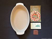 Top view of beige stoneware oval baker with scraper, care card and recipe booklet