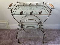 Front view of metal bar cart showing two glass shelves, leaf accents, wood handles, lucite wheels