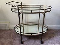 Full side view of oval brass-tone two-tier glass bar cart on carpeted floor