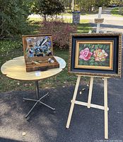Group view of framed painting on easel beside paint box