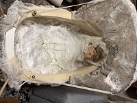 Top view of porcelain doll lying inside lace-lined wicker pram
