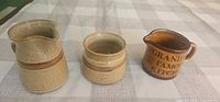 Three small ceramic vessels grouped: beige stoneware pitcher, matching jar, brown text pitcher