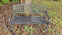 Front view of slatted metal garden bench under tree showing full structure and wear