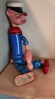 Side view of wooden Popeye doll seated on shelf showing overall form, paint colors, jointed limbs, and wear