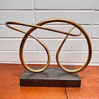 Front view of gold infinity loop metal sculpture on black base against white brick background