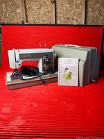 Sewing machine on base with foot pedal, detached case lid and instruction manual in front of red backdrop