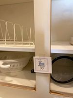cabinet shelf showing white metal plate rack, partial view of colander and black mesh strainer