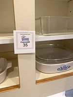 View of cabinet shelves with clear glass rectangular baking dish on upper shelf and blue Cornflower CorningWare casserole with lid on lower shelf