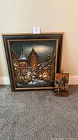 Square and rectangular framed village scene paintings leaning against wall