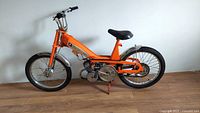 Left side view of orange 1971 Motobecane moped showing frame, engine, pedals and fenders