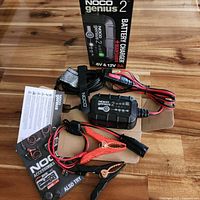 Full lot showing NOCO Genius 2 charger, clamps, cables, manual, damaged box