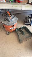 Ridgid shop vacuum with power cord on lid next to large green dust pan