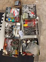 Top-down view of six open metal tins containing assorted drill bits, wires, fasteners, electrical components, and a small cardboard box