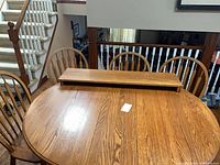 Oval oak dining table with leaves stacked on top and four Windsor chairs in background