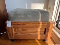 Front angled view of wooden ottoman with sage green cushion and caster wheels