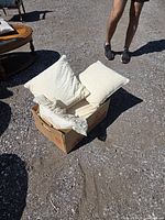 Cardboard box containing five white/beige throw cushions outdoors on gravel