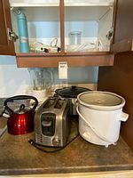 Red kettle, stainless toaster, white slow cooker, blender base and jar, partial view of cabinet contents